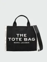 Bolso tipo tote bag de Marc Jacobs en color beige, confeccionado en lona resistente. Presenta el texto "THE TOTE BAG" y "MARC JACOBS" impreso en la parte frontal en color negro. Cuenta con asas superiores y una correa de hombro ajustable y extraíble, además de ribetes laterales negros con inscripciones.