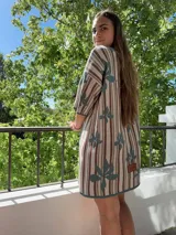 Vestido tejido de hilo de algodón con diseño de rayas verticales en tonos beige, marrón y verde, y estampado de hojas verdes. Tiene manga corta, cuello redondo y silueta holgada.