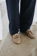 Mocasín estilo náutico de gamuza marrón, con cordones de cuero, borla decorativa y cuentas de colores en el empeine.