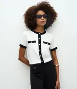 Cardigan de punto de manga corta con cuello redondo y cierre frontal de botones perolados. Presenta un diseño bicolor en blanco y negro con detalles de contraste en cuello, puños, tapetas de bolsillos frontales y vista central.