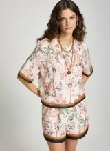 Blusa estampada Le Lis Blanc con diseño de paisaje tropical y arquitectónico, cuello redondo y mangas cortas con ribetes en tonos marrón y negro.