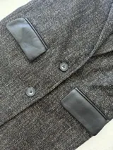 Tapado de lana en color gris jaspeado, con solapa clásica y cierre frontal de botones. Presenta detalles en cuero negro en las tapas de los bolsillos.