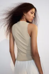 Top de algodón color beige con escote en V, sin mangas y cierre frontal con botones. Presenta un diseño asimétrico en el bajo con abertura delantera.