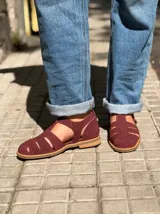 Sandalia tipo cangrejera de gamuza color bordó, con diseño cerrado de tiras cruzadas, talón cerrado y tira con hebilla metálica ajustable en el tobillo.