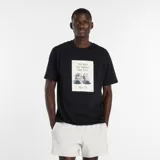 Remera de algodón color negro, de corte clásico y manga corta. Presenta un estampado frontal rectangular con la imagen de un par de championes New Balance 574 y el texto 'The Most New Balance Shoe Ever. The 574'.