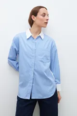 Camisa celeste con rayas verticales blancas, de corte amplio y cuello camisero clásico. Presenta bolsillo de parche en el pecho, espalda lisa en color blanco y puños abotonados.