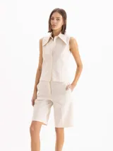Camisa sin mangas color beige, de calce entallado y largo hasta la cadera, con cuello clásico y cierre frontal de botones.
