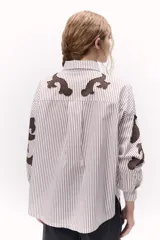 Camisa a rayas verticales blancas y marrones con bordados localizados y apliques metálicos. Tiene mangas abullonadas y cuello clásico con broche metálico.