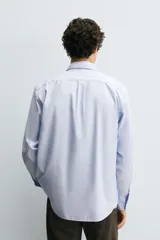 Camisa de algodón de corte regular, color celeste claro con finas rayas blancas verticales. Presenta cuello abotonado (button-down), manga larga con puños abotonados y un bolsillo plastrón en el pecho izquierdo.