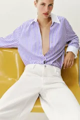 Camisa de popelín con diseño de rayas verticales en tonos blanco y lila, cuello solapa con escote en pico, manga larga y puños anchos contrastantes en color blanco. Cierre frontal con botones metálicos dorados.