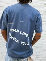 Remera oversize de algodón con efecto lavado, color azul oscuro. Presenta un estampado minimalista en blanco en el pecho con texto en dos líneas: "URBAN LIFESTYL" y "RAQRA YTIJAU" (lectura invertida de "QUALITY APPAREL"), y la palabra "OSTEN" en el lado derecho.