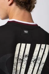 Camiseta de estilo deportivo inspirada en el fútbol americano, con diseño de bloques de color en negro y blanco. Presenta cuello en V con detalles a rayas, mangas cortas anchas y estampado frontal con efecto desgastado que incluye gráficos y texto.