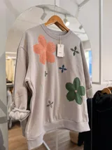 Buzo de cuello redondo en color beige, con diseño oversize y felpa interna. Presenta bordados de flores en tonos rosa, verde y azul distribuidos en el frente.