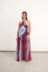 Vestido largo sin mangas, con escote halter y cuello alto ajustable. Presenta un diseño combinado con estampa floral grande en la parte superior y falda larga con patrón de rayas verticales en tonos bordó, celeste y blanco.