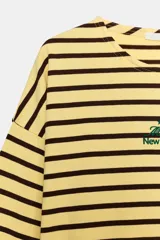 Camiseta oversize de manga larga con diseño de rayas horizontales amarillas y negras. Presenta cuello redondo y un detalle de texto bordado en el centro del pecho.
