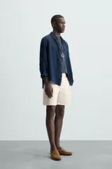 Bermuda de hombre color beige claro, corte regular fit, con pliegues frontales, bolsillos delanteros y bolsillos traseros de doble vivo con botón.