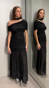Vestido largo negro ajustado con diseño drapeado en todo el cuerpo, un hombro descubierto, cuello tipo choker integrado y falda larga semitransparente con vuelo en el inferior.