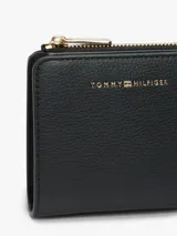 Billetera compacta Tommy Hilfiger de diseño cuadrado, confeccionada en material sintético con textura granulada. Cuenta con cierre perimetral metálico dorado y logo de la marca aplicado en letras metálicas doradas en el frente.