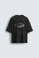Camiseta de punto de algodón, corte boxy fit, color negro con efecto lavado. Presenta un estampado gráfico frontal que simula un ojo o forma ovalada con texto ilegible en su interior.