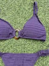 Conjunto de bikini color violeta confeccionado en tela texturada. El corpiño es de estilo top con una hebilla ovalada marrón en el centro y breteles finos. La bombacha es de corte colaless con detalle de frunces en los laterales.