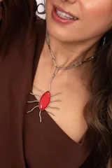Collar con cadena de eslabones metálicos y un gran dije colgante con forma de sol o araña, que presenta una piedra de cristal rojo en el centro y patas metálicas onduladas.