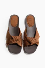 Sandalias estilo zueco color marrón, con tiras de gamuza anudadas en la parte delantera y detalles de tachas metálicas doradas en los laterales. Cuenta con suela de madera oscura de tacón cuadrado bajo.