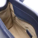 Cartera tipo tote pequeña de textura denim azul con detalles en cuerina azul oscuro, asas de mano y correa de hombro ajustable. Cuenta con un bolsillo frontal con cierre y el logo de la marca impreso.