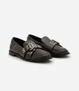 Mocasín femenino de cuero sintético color marrón oscuro, con punta redondeada y suela plana. Presenta un diseño con tiras cruzadas en el empeine decoradas con ojales metálicos y una hebilla ajustable.