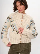 Cárdigan tejido a mano color crudo, con diseño de trenzas y flores bordadas en azul y marrón. Tiene cuello redondo y cierre frontal con botones de madera.