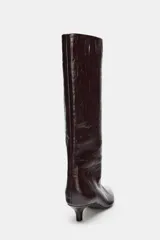 Bota de caña alta con acabado efecto craquelado en color marrón oscuro. Presenta un diseño de punta fina, tacón kitten y una estructura de calce fácil tipo pull-on.