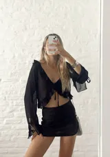 Blusa corta negra de mangas largas acampanadas, con volados en el escote y lazo ajustable en el frente.