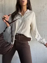 Camisa de color beige con cuello clásico, cierre frontal de botones y detalle de bordado geométrico en el pecho. Presenta mangas largas con puños y un diseño de tela con textura ligera.