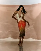 Vestido corto de crochet color marrón con acabado brillante, diseño ajustado al cuerpo, escote halter con tirantes de cadena metálica dorada y abertura frontal pronunciada.