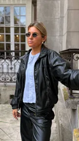 Campera de cuero sintético color negro, con cuello camisero, cierre frontal y costuras visibles en contraste. Presenta un diseño holgado con puños elásticos.