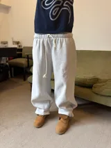 Pantalón de jogging gris melange de corte baggy, confeccionado en algodón. Presenta cintura elástica con cordón largo ajustable, bordado decorativo en la pierna izquierda y botones metálicos en la parte inferior de la botamanga para ajustar el ancho.