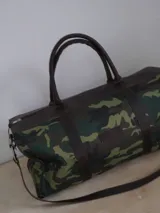 Bolso de viaje tipo duffel de lona negra con detalles en cuero tostado, asas de mano y correa ajustable para el hombro.