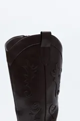 Bota de caña alta estilo western confeccionada en cuero sintético, con bordados decorativos en relieve a lo largo de la caña y puntera afinada.