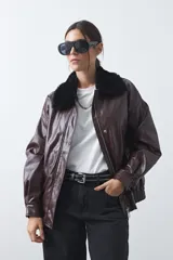 Campera de cuero sintético color bordó con acabado satinado, corte relajado y elástico en la cintura. Presenta cuello de piel sintética negra, cierre frontal con cremallera y botones a presión.
