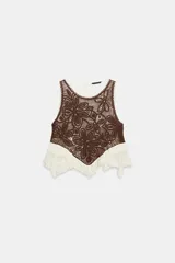 Top sin mangas con cuello redondo, confeccionado en una combinación de tejido de crochet marrón con diseño floral y base de tela color crudo con volados asimétricos y detalles de puntilla. Cuenta con cierre de botón en la espalda.