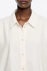 Camisa de corte recto en color blanco, con cuello clásico y cierre frontal de botones. Presenta mangas largas con puños abotonados y una textura sutil en el tejido.