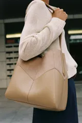 Bolso de hombro color tostado con diseño estructurado que combina paneles de cuero liso y gamuza con pespuntes visibles. Cuenta con un asa larga de hombro ajustable y cierre superior con botón imantado.