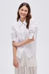 Camisa blanca de algodón, holgada, de manga larga con puños anchos, cuello camisero y cierre frontal oculto con un botón dorado visible en el cuello.