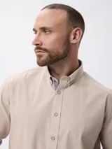 Camisa de manga larga en algodón tramado, con cuello clásico abotonado y cierre frontal con botones. Presenta un diseño liso con textura visible y un pequeño bordado en la pechera.