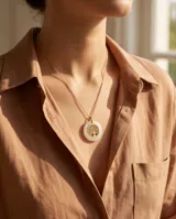 Collar de acero con baño de oro, compuesto por una cadena fina y un dije circular que presenta un árbol de la vida dorado en el centro, enmarcado por un borde de cerámica blanca.