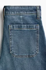 Jean de tiro alto con corte wide leg, confeccionado en denim de algodón con leve elasticidad. Presenta bolsillos de parche frontales, cierre con botón y costuras marcadas a lo largo de las piernas.