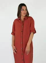 Vestido largo de lino marrón con cierre frontal de botones, cuello camisero, mangas tres cuartos con presilla y costuras decorativas blancas en la botonera.