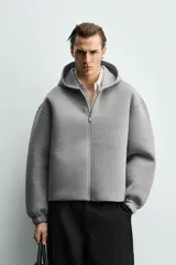 Sudadera gris de corte boxy fit con capucha y cierre frontal de cremallera. Confeccionada en tejido doble y compacto, cuenta con bolsillos laterales ocultos en la cadera y mangas largas con puños elásticos.