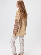Pullover de tejido acanalado grueso con cuello alto y cierre frontal parcial. Presenta un diseño de bloques de color, con cuerpo en tono beige y mangas en contraste con rayas verticales en tonos rojizos. Incluye un detalle bordado en el pecho.