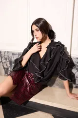 Blusa fluida color negro de corte holgado, con cuello alto, mangas largas y llamativos volados en cascada que recorren el frente y las mangas, decorados con costuras en hilo blanco en los bordes.