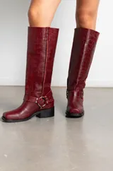 Botas de caña alta en cuero con textura de cocodrilo, color bordó. Presentan un diseño de punta cuadrada, taco bajo y una correa decorativa con argolla metálica y remaches en la zona del tobillo.